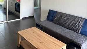 A modern living room with a wooden coffee table, a grey sofa with blue cushions, and a glass door leading to a kitchen. - Trésor Caché d'Amazonie Oasis de Calme (Cayenne)