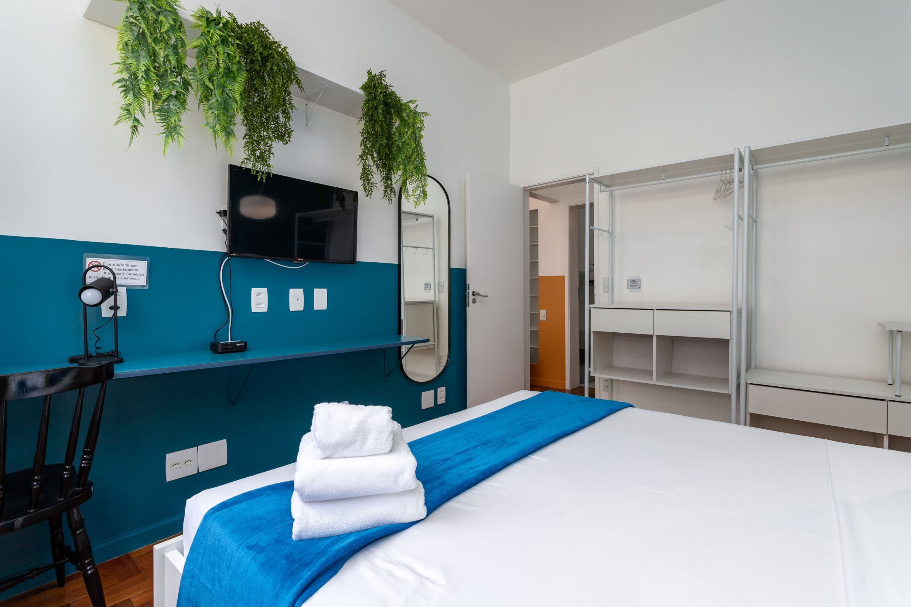 A hotel room with a bed, a desk with a chair, a TV, a mirror, and a wardrobe.