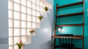 A room with a teal accent wall, a wooden ladder shelf, a desk with a chair, and a wall-mounted shelf with plants. - For Families 10 Minutes From Ipanema Beach Ns103 (Rio de Janeiro)