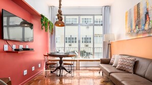 A modern living room with a red accent wall, a wooden floor, a sofa with patterned cushions, a dining table with chairs, and a large window with a view of buildings. - For Families 10 Minutes From Ipanema Beach Ns103 (Rio de Janeiro)