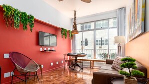 A modern living room with a red accent wall, a wooden herringbone floor, a dining area with a round table, and a large window offering a view of buildings. - For Families 10 Minutes From Ipanema Beach Ns103 (Rio de Janeiro)