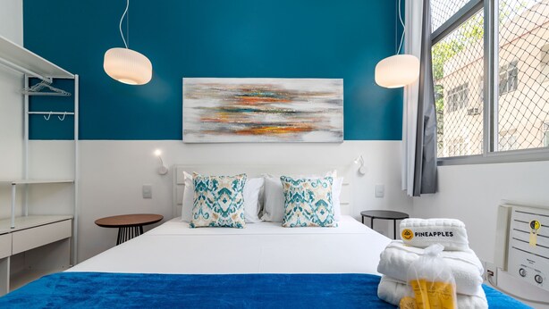 A modern bedroom with a large bed, two pillows, a blue blanket, a painting on the wall, and a window with a view. - For Families 10 Minutes From Ipanema Beach Ns103 (Rio de Janeiro)