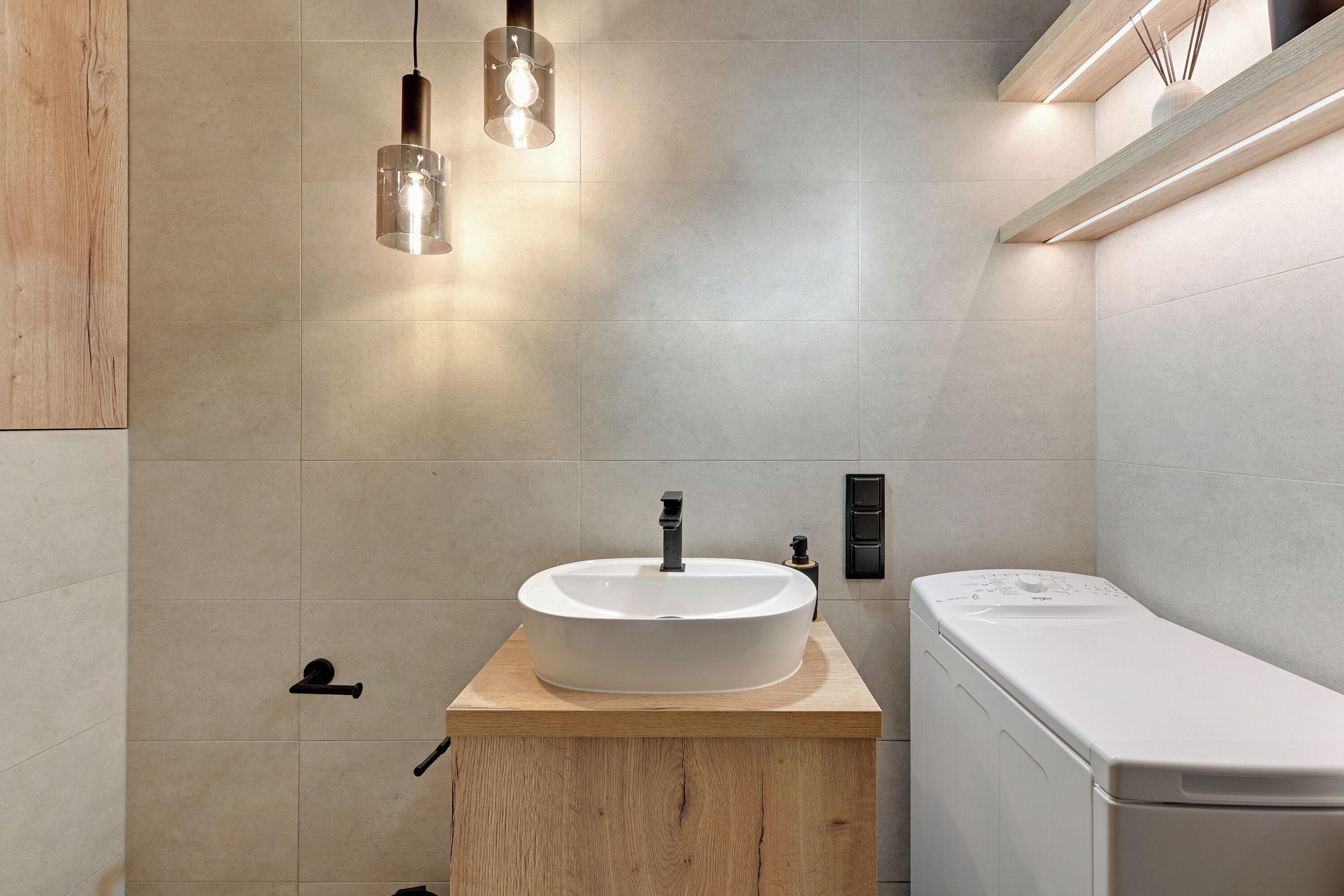 Een moderne badkamer met een houten wastafelmeubel, een witte wasbak en een toilet. Boven de wasbak hangen pendelverlichting en er is een muurbevestigde zeepdispenser.