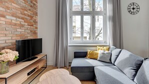A modern living room with a brick wall, a flat-screen TV on a wooden stand, a grey sofa with cushions, a wooden coffee table, and a window with curtains. - Forma by Grand Apartments (Sopot)