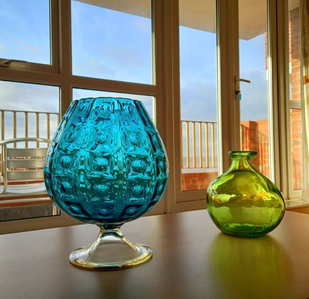 Deux vases en verre sont posés sur une table près d’une fenêtre, d’où l’on peut admirer des bâtiments.