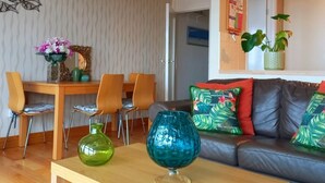 Ett modernt vardagsrum med ett soffbord i trä, en soffa med färgglada kuddar, en matplats med ett bord och stolar i trä samt en vägg med ett dekorativt konstverk föreställande ett spindelnät.