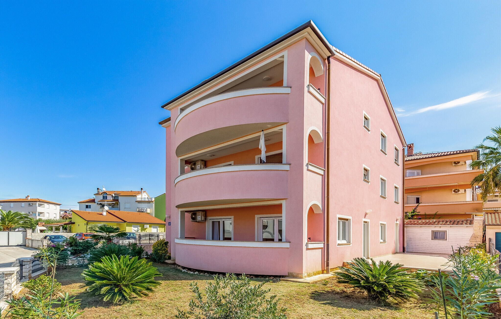 Un immeuble rose à plusieurs étages, doté de balcons, un parking avec des voitures et une végétation luxuriante tout autour.