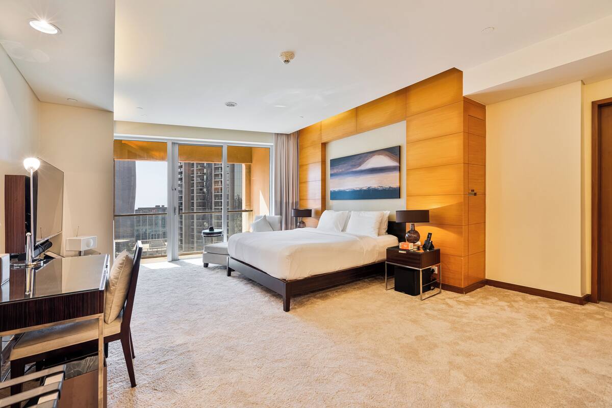 A modern hotel room with a large bed, a desk with a chair, a small table, and a large window offering a city view.
