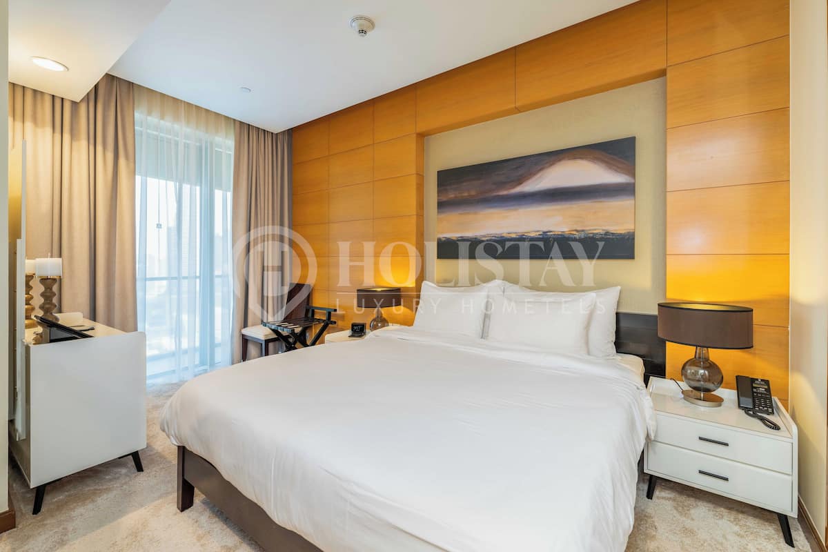 A luxury hotel bedroom with a large bed, a desk, a chair, a nightstand with a lamp, and a painting on the wall.