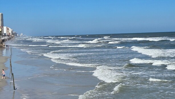 A beach with waves, a clear sky, and buildings in the background.