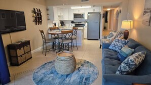 A modern living room with a blue sofa, a coffee table, and a TV. There is a kitchen in the background with white cabinets and a refrigerator.