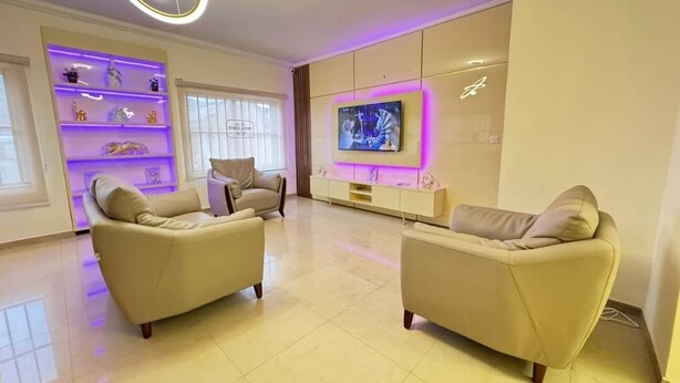 Una sala de estar moderna con dos sofás color beige, un televisor montado en la pared y una estantería iluminada con luz púrpura.