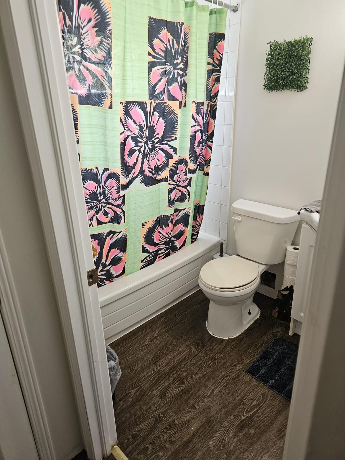 Ein Badezimmer mit Toilette, einer Duschvorhang mit Blumenmuster, einer kleinen grünen Pflanze an der Wand und einem dunklen Holzboden.
