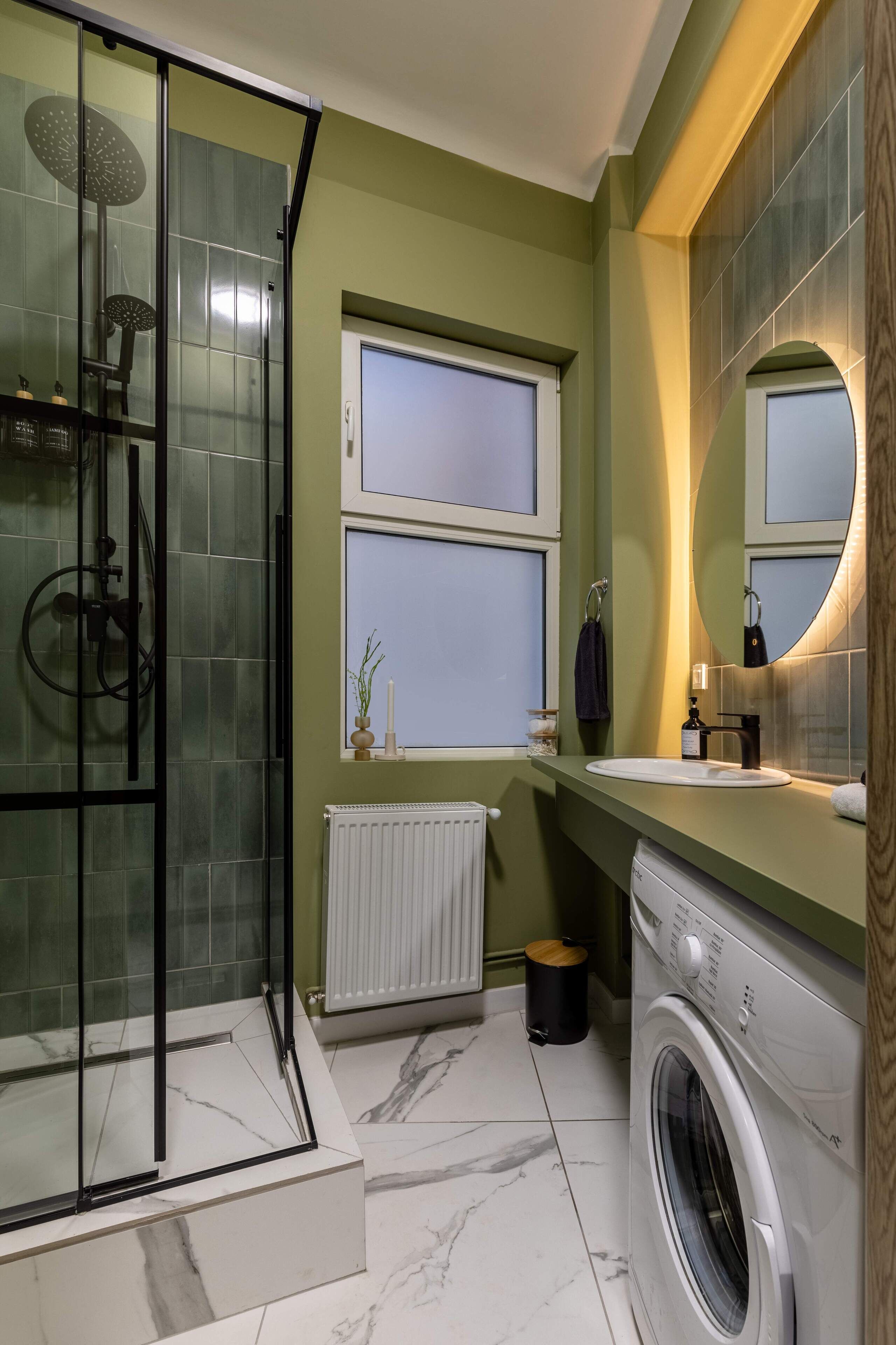 A modern bathroom with a glass shower enclosure, a built-in washing machine, and a window with a view.
