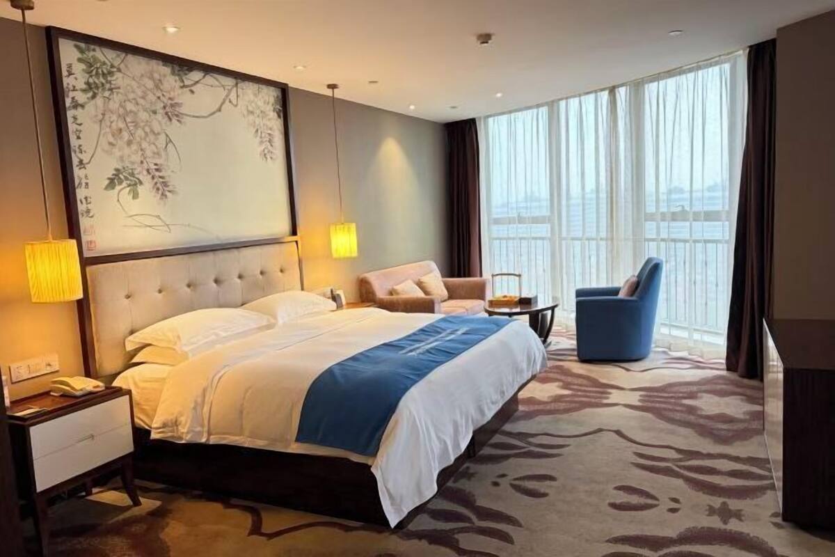 Habitación de hotel con una cama grande, una silla, una mesita de noche, una lámpara y vistas al exterior.