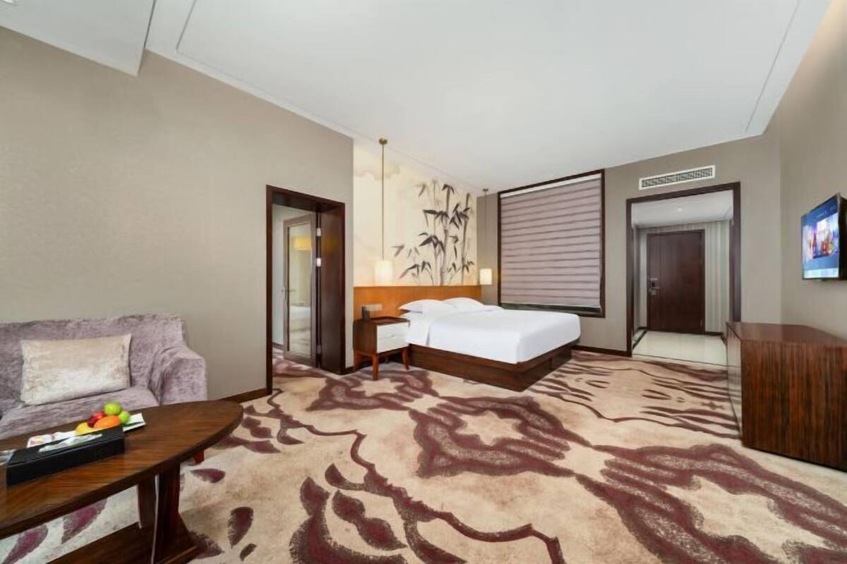 Habitación de hotel con cama, sofá, escritorio, una pared decorada con una obra de arte de bambú y una alfombra estampada.