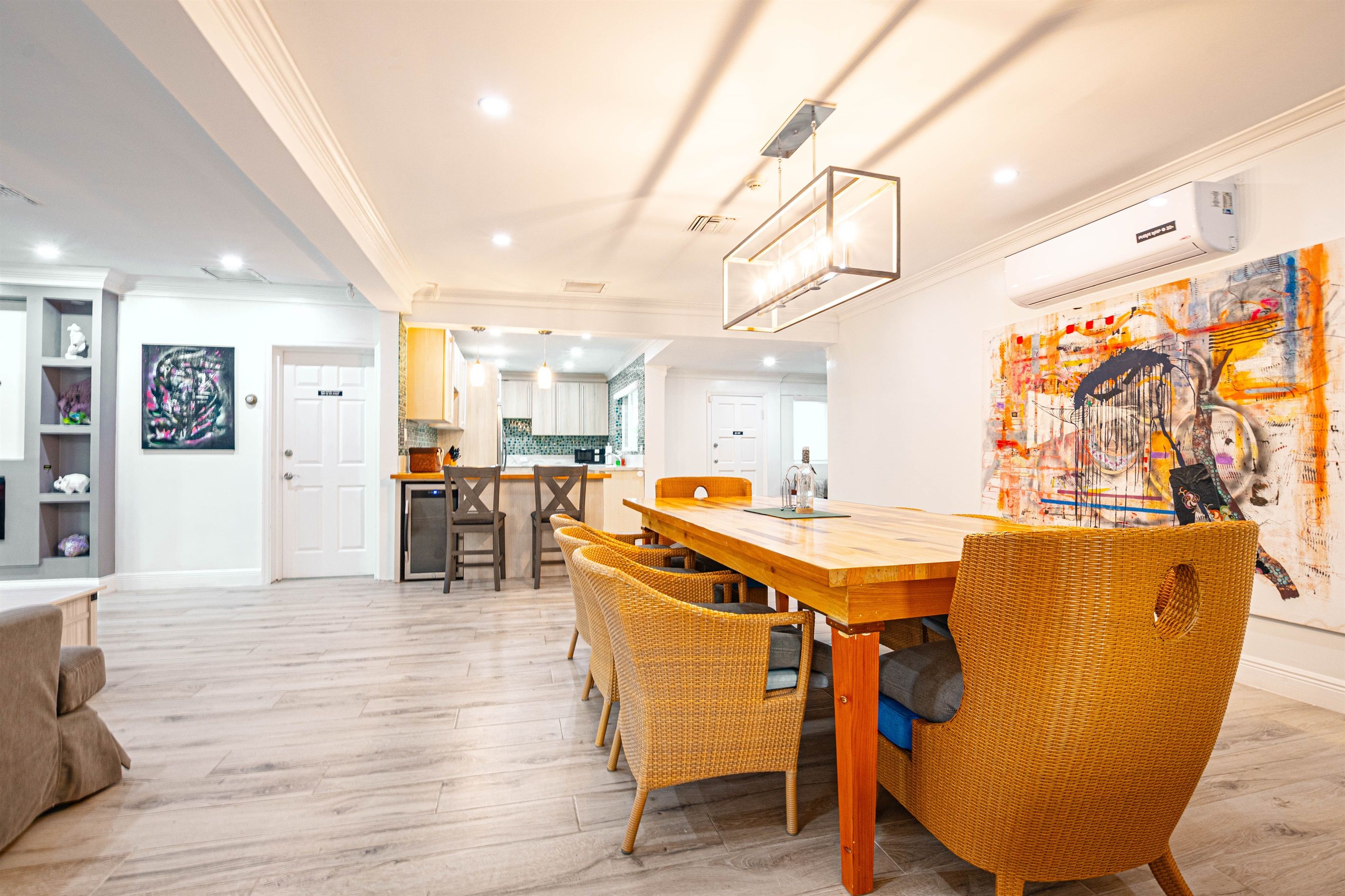 Ein modernes Interieur mit Essbereich, der einen Holztisch und Korbstühle umfasst, einer Küche mit weißen Schränken sowie einem Wohnzimmer mit einer Couch und einem großen abstrakten Gemälde an der Wand.