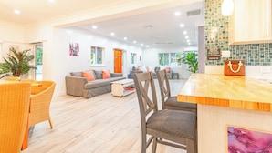 Amplia sala de estar con una mesa de comedor de madera, un sofá y sillas. Se observa una zona de cocina con salpicadero de azulejos y encimera de madera.