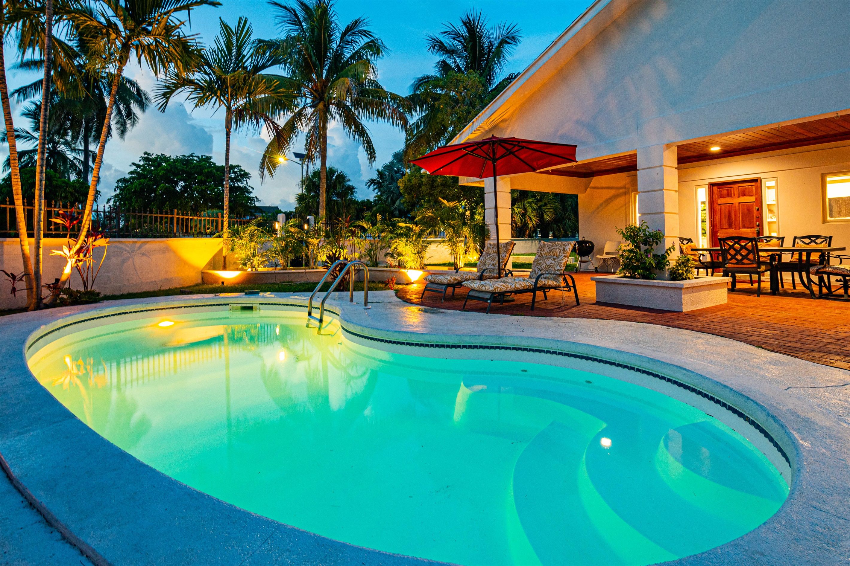 A swimming pool with lounge chairs and a red umbrella, surrounded by palm trees and a building with a patio.