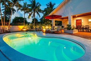 Une piscine avec des chaises longues et un parasol rouge, entourée de palmiers et d’un bâtiment doté d’une terrasse.