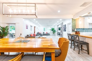 Un espace de repas moderne avec une table en bois et des chaises en osier, une cuisine avec un dosseret carrelé et un salon avec un canapé et un téléviseur.