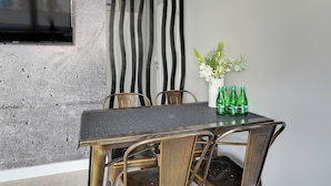 Un coin repas comprenant une table, des chaises, un téléviseur fixé à un mur texturé, ainsi qu’un vase avec des fleurs et des bouteilles vertes.