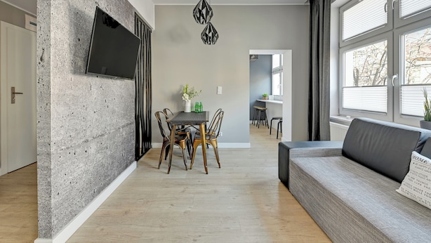 Un salon moderne avec un canapé gris, une table à manger avec des chaises, un téléviseur à écran plat fixé à un mur texturé et une fenêtre avec des stores.