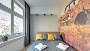 Un lit bien fait, recouvert d’une courtepointe grise matelassée, avec deux oreillers jaune et gris, et deux serviettes blanches pliées posées sur le lit.