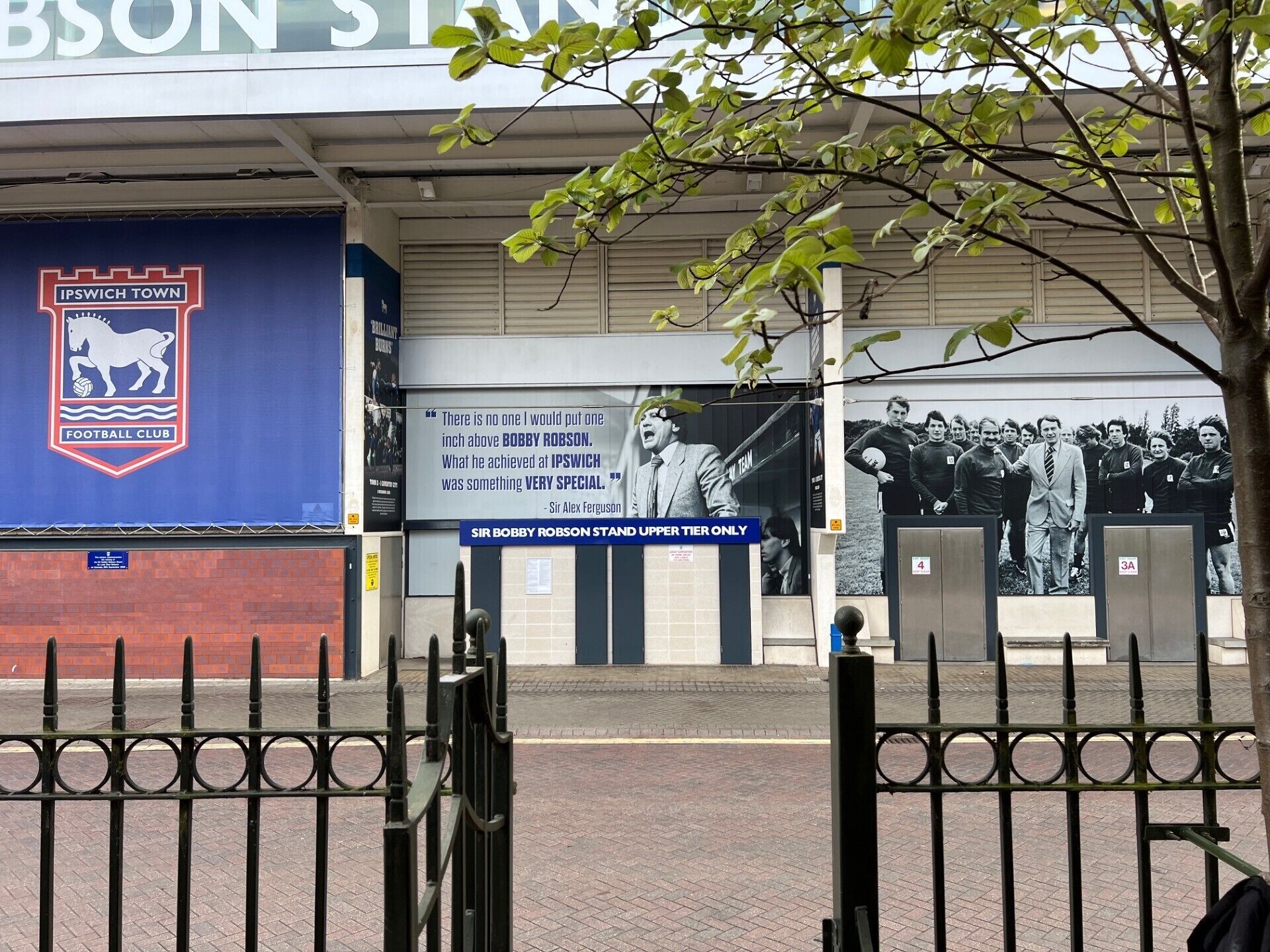Entrada a un club deportivo con pared azul, un logotipo de caballo y una cita sobre Sir Bobby Robson.