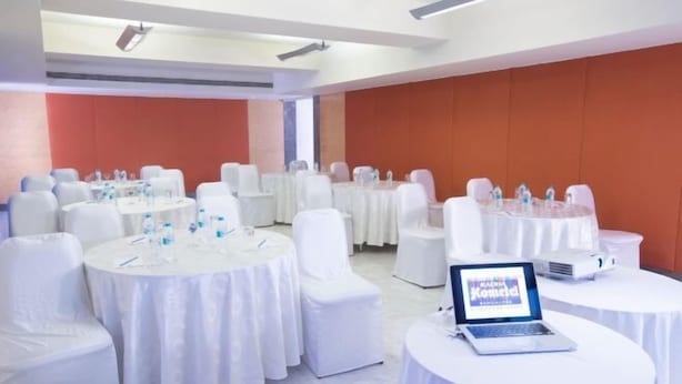 A conference room with multiple round tables, each covered with white tablecloths and equipped with chairs. There are laptops on some tables, and water bottles are visible. The room has a modern design with white and orange walls. - Radha Hometel Bengaluru (Bengaluru)
