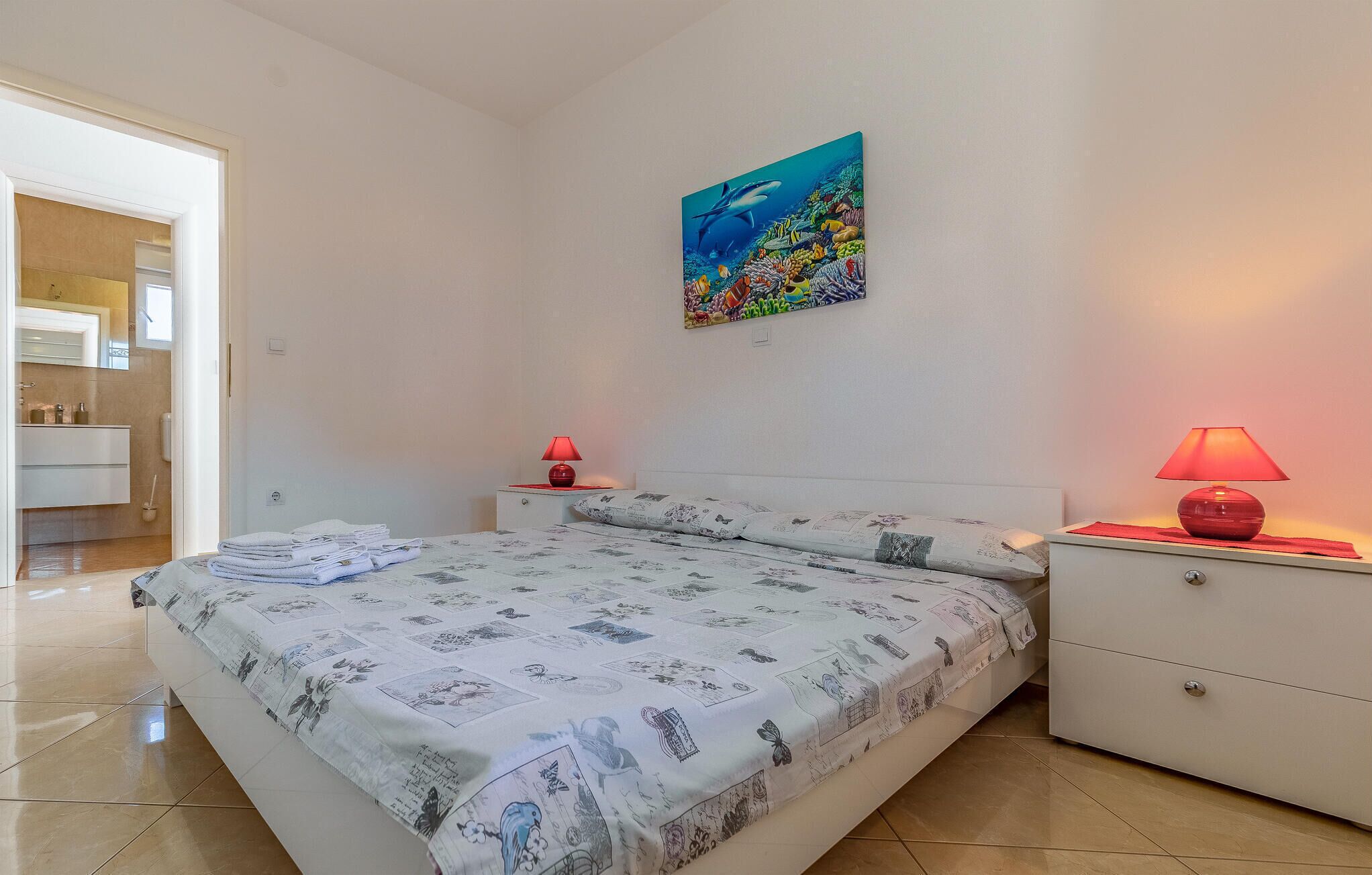 Une chambre à coucher avec un lit, deux tables de chevet, une commode, un tableau représentant une scène sous-marine, et une salle de bain visible à travers une porte ouverte.