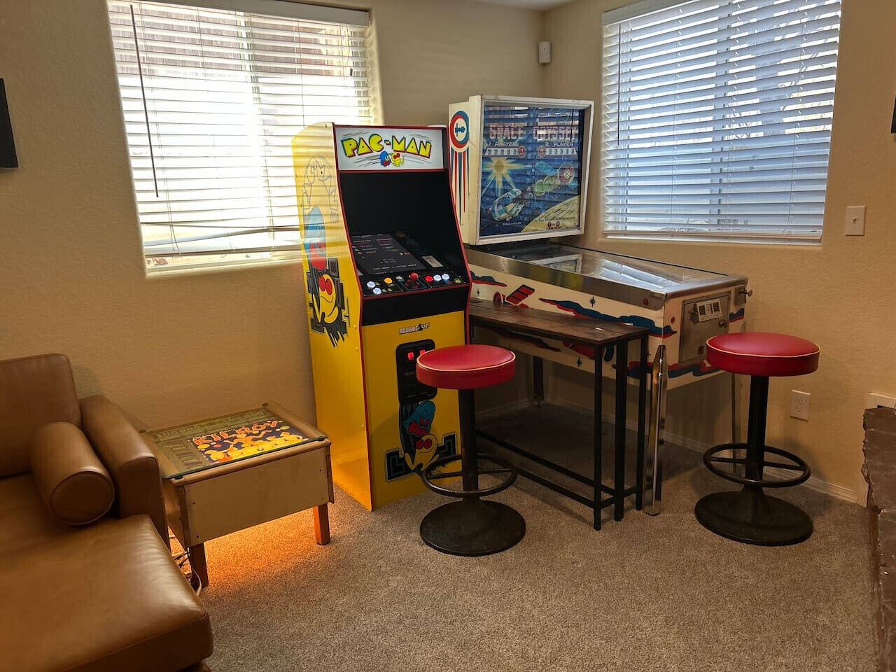 Een kamer met een Pac-Man spelcomputer, twee rode barkrukken, een bruine leren fauteuil en een kleine tafel met een puzzel.