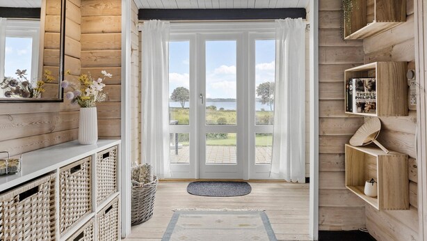 A wooden interior with a large glass door leading to a view of a lake and trees. There are wicker baskets, a white vase with flowers, and a shelf with books and a decorative item. - Stunning home in Aabenraa (Aabenraa)