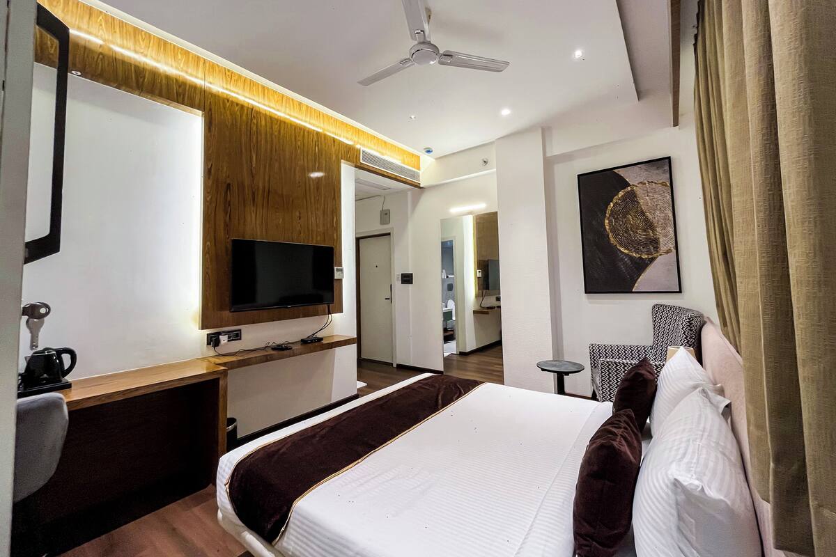 Una habitación de hotel moderna con un televisor de pantalla plana, un tocador de madera, una cama con edredón blanco y marrón, un ventilador de techo y un cuadro en la pared.