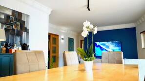 A dining area with a wooden table, white chairs, and a vase with white flowers. There is a television on the wall and a kitchen area in the background. - Spacious Penthouse w/ Barbecue, wifi, 2 terraces & Views (Praia)