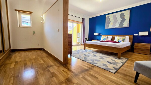 A bedroom with a wooden floor, a bed with a patterned rug, a blue accent wall, and a window with curtains. - Spacious Penthouse w/ Barbecue, wifi, 2 terraces & Views (Praia)