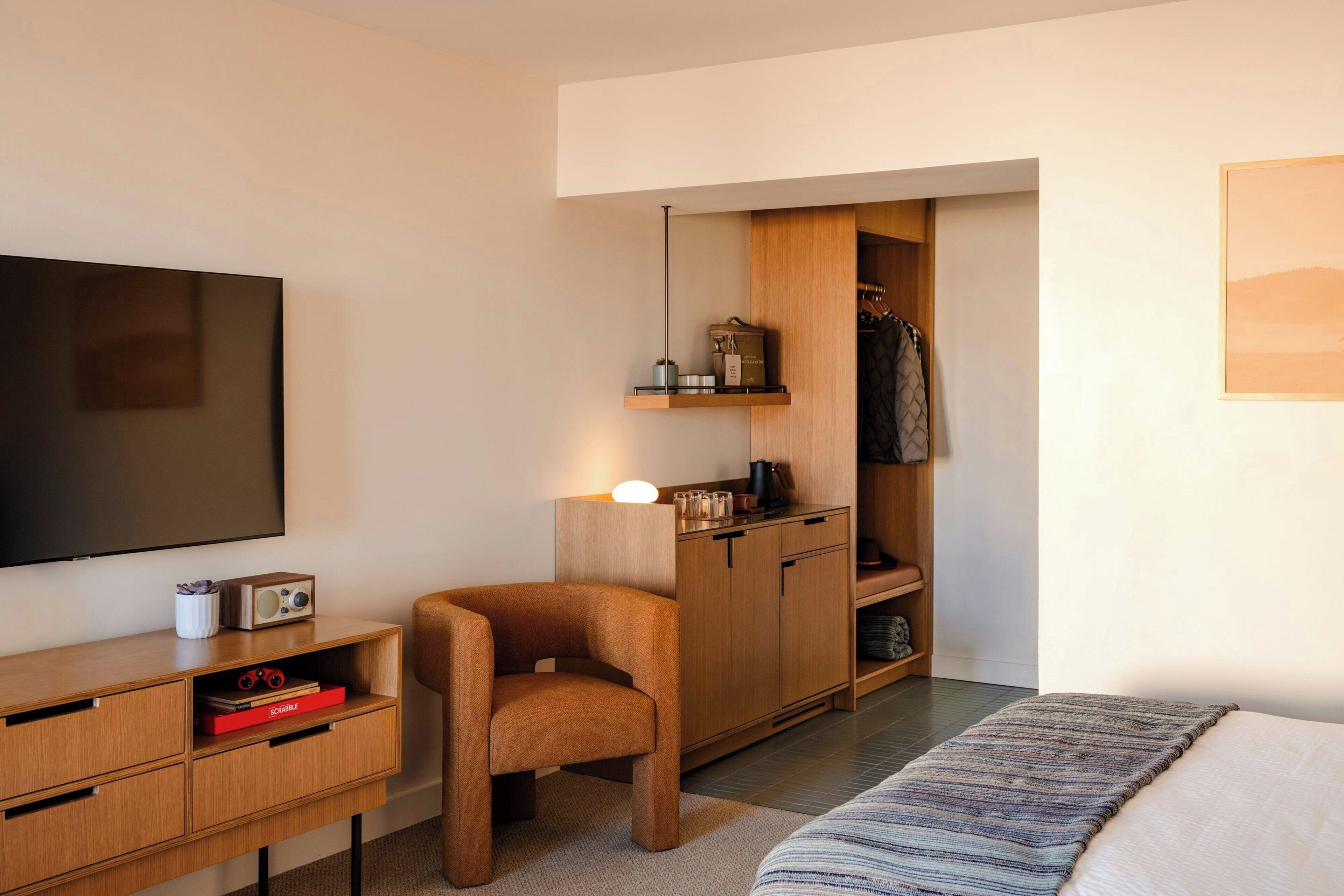 A compact hotel room with a bed, a TV, a wooden desk, a chair, a wardrobe, and a shelf with items.