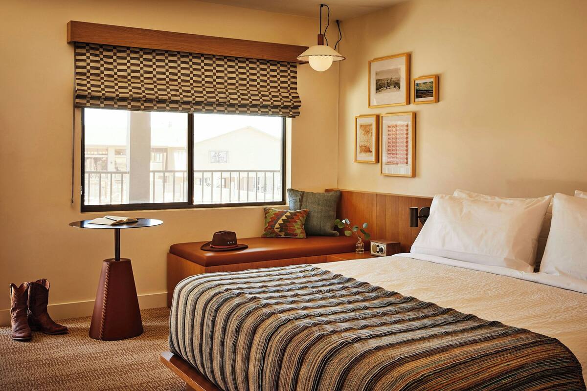 Une chambre d’hôtel avec un grand lit, une petite table ronde, un banc et une fenêtre avec un store à carreaux.