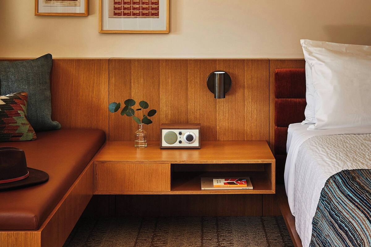 Une chambre d’hôtel avec une tête de lit en bois, une table de chevet avec une radio, une petite étagère et un lit avec du linge de lit blanc.