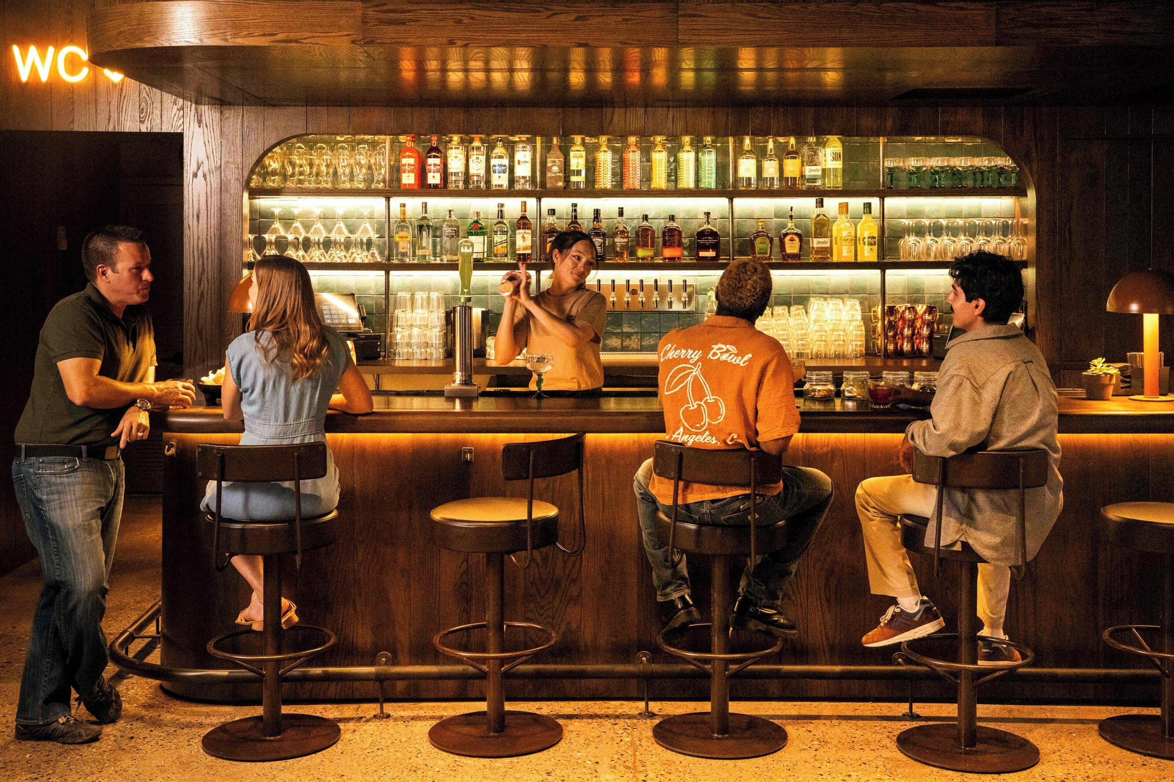 Zona de bar con taburetes, un bartender y una variedad de botellas y vasos en estantes.