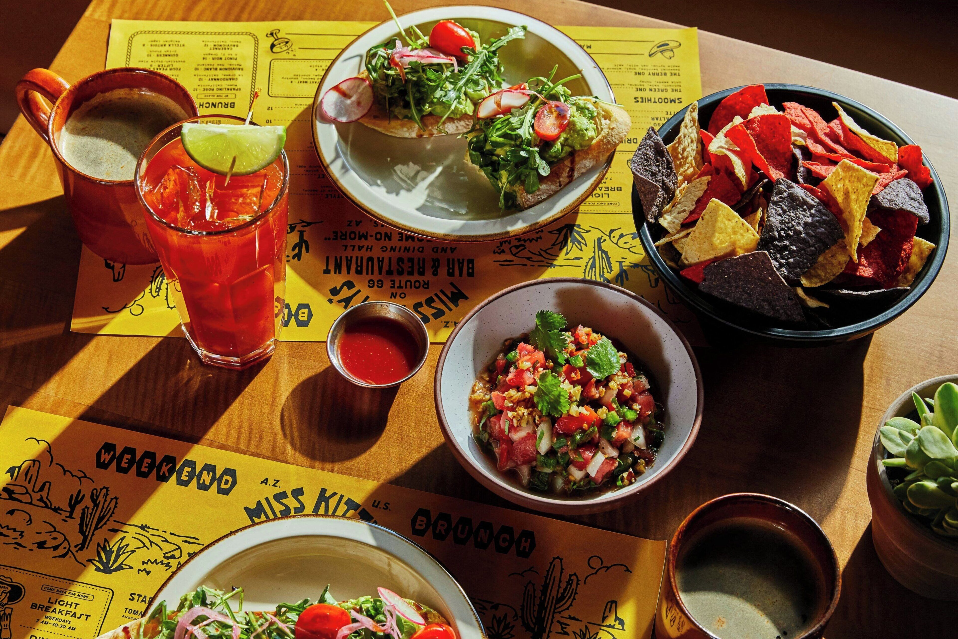 A table with a variety of dishes including a salad, a bowl of chips, a drink, and a small bowl of salsa.