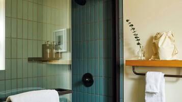 A bathroom with a shower, a sink, and a mirror. There is a towel hanging on a rack and a shelf with toiletries.