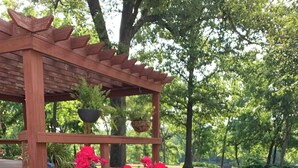 Una pergola in legno con una panchina, circondata da un prato verdeggiante e alberi.