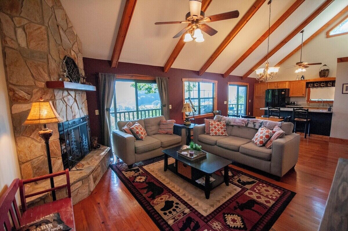A cozy living room with a stone fireplace, a large sofa set, a coffee table, and a kitchen in the background.