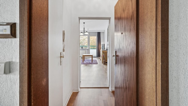 Un pasillo con puertas de madera, una pared blanca y vistas a una habitación con ventana, un banco y una mesa.