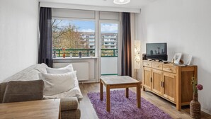 Una sala de estar moderna con un sofá, una mesa de madera, un televisor sobre un mueble de madera y vistas de edificios a través de amplios ventanales.