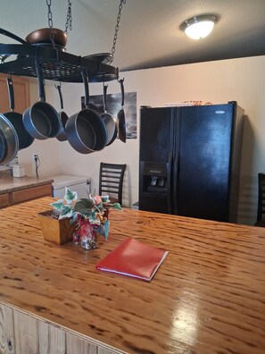 Una cocina con ollas colgantes, un refrigerador, una isla de madera con un florero y una libreta.