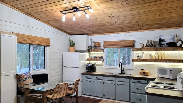 A kitchen with wooden ceiling, white walls, and grey cabinets. There is a dining table with chairs, a refrigerator, a microwave, and a sink. - Pinescape: Cabin•WiFi•Wood Fireplace•Pet friendly•Fish•Relaxation (Big River)