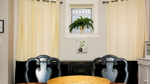A dining area with a wooden table, two chairs, a potted plant on the window sill, and a white cabinet to the right.