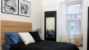 A bedroom with a bed, a window with blinds, a mirror, and two framed abstract artworks on the wall.
